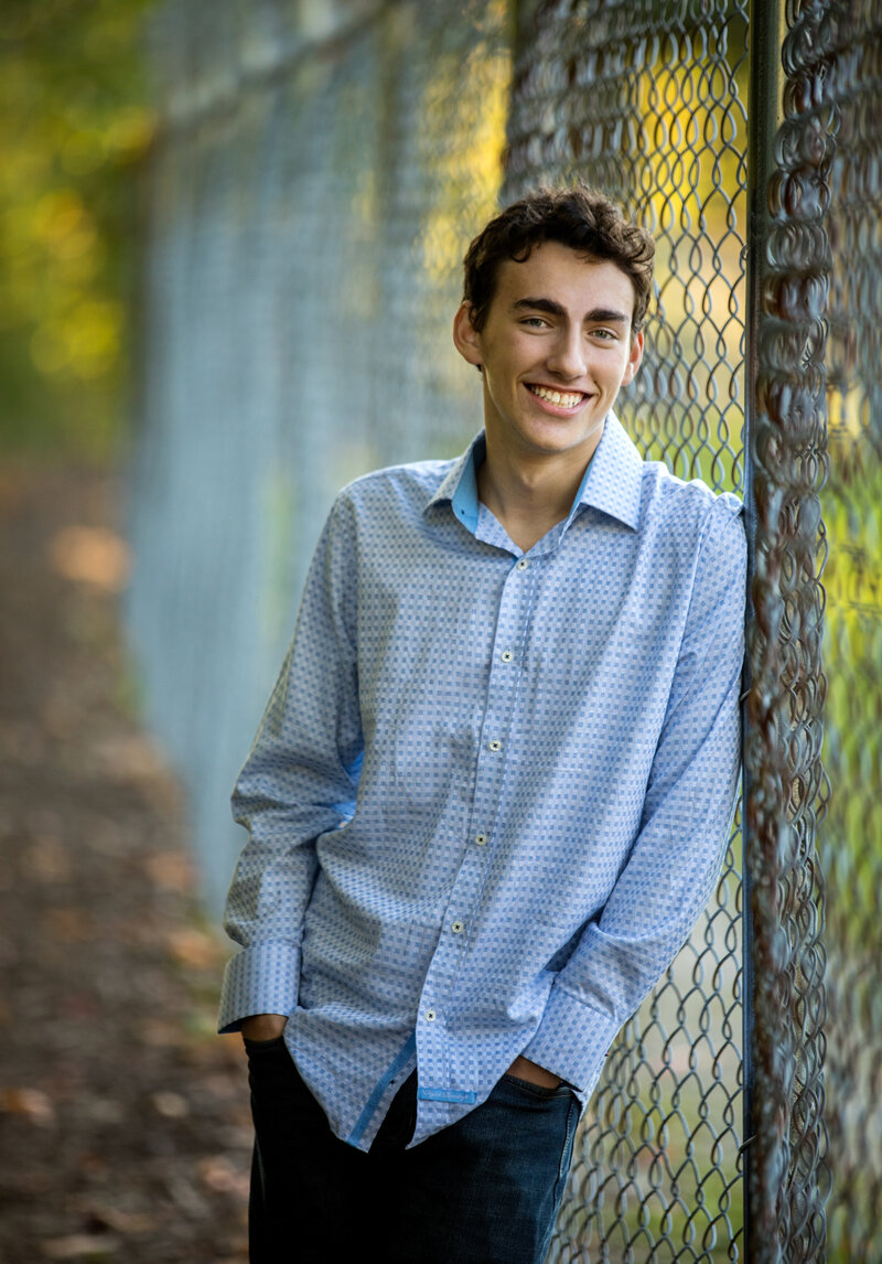 A senior boy wears a dress shirt and leans against a fence.