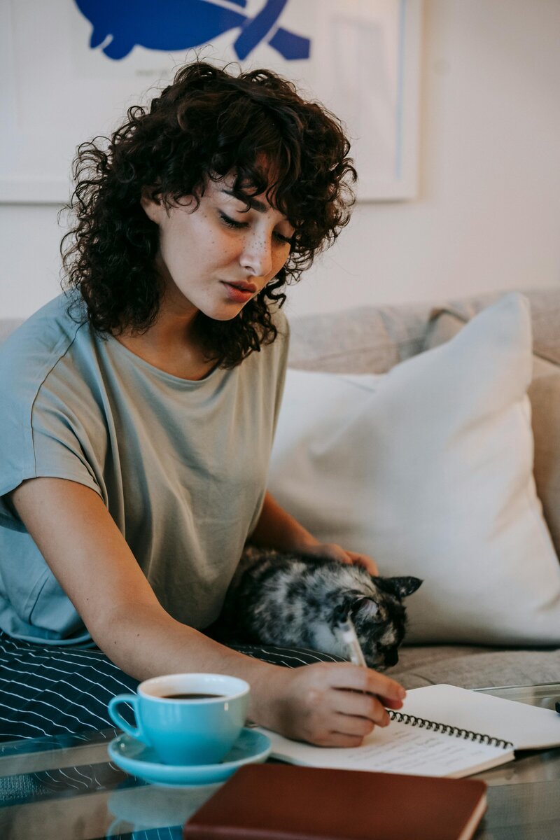 a photo of a woman writing in a notebook on her couch with a cup of coffee next to her and petting a cat that is sitting next to her