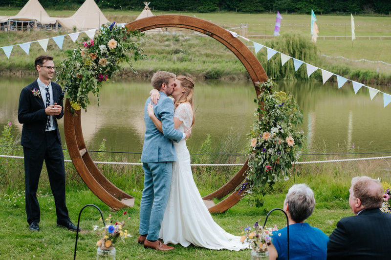 Bride and groom embrace and take their first kiss as man and wife, in front of a lake at their Hadsham farm wedding.  A large metal flower ring and pastel coloured bunting decorates the ceremony space behind them