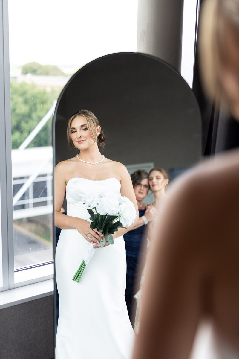 This is a photo of a bride and Mother of bride and sister in a mirror reflection. - The bride is wearing a white strapless dress and holding a bouquet of white roses. - The Mother of the bride is wearing a dark blue dress and standing behind the bride. - The background shows a view of trees and a building through a window. - The photo is taken from the perspective of someone standing in front of the mirror.