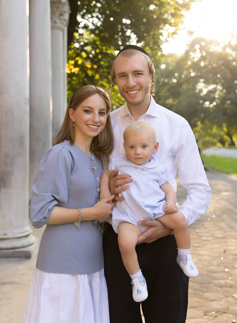 Mom, dad, and baby boy pose for a first birthday family photoshoot at golden hour in a brooklyn, NY park. There is a stone portico behind them with soft lift. All are standing and looking at the camera, smiling. Captured by premier Brooklyn NY family photographer Chaya Bornstein Photography.