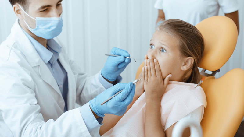 A little girl holds her hands over her mouth in fear, so that the dentist can't work on her mouth.