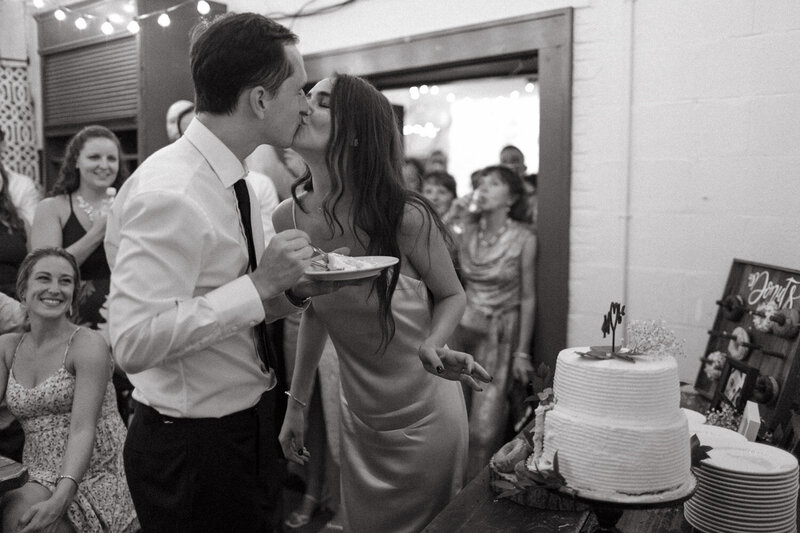 A couple kissing after cutting their wedding cake while all of their family cheer them on.