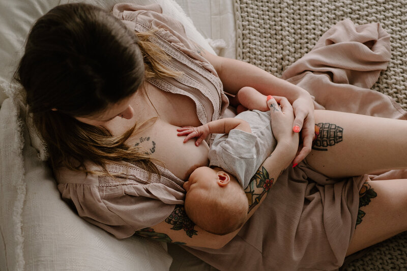 Breastfeeding mom in beige laying on bed in lifestyle studio with baby girl in gray.
