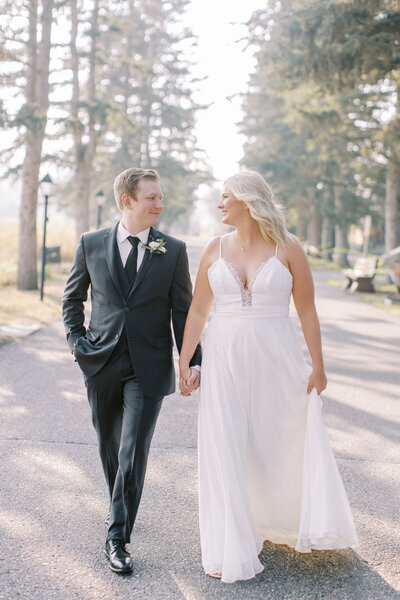 A couple in wedding attire walk hand in hand on a tree-lined path, smiling at each other, their joy guided by the expertise of a full-service wedding planner.