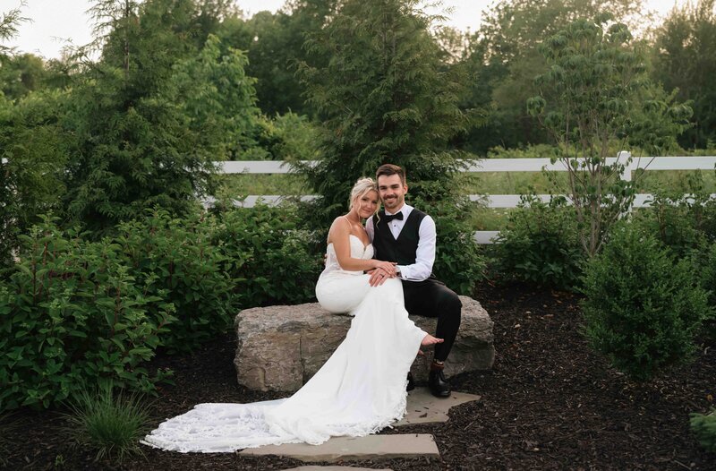 A bride and groom sit snuggling on a large rock outside at their summer wedding at golden hour