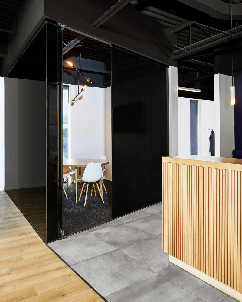A meeting room with opaque black glass walls and a small wood table and modern chairs inside next to a tall wood paneled reception desk