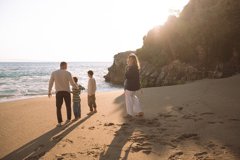 Family walking barefoot on the sand at Laguna Beach during golden hour, parents and children looking out at the ocean with warm candid light — Orange County storyteller session by Maria Alcantara Photography