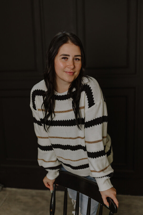 Britt posing for a portrait inside of a studio with black walls. She's leaning against a black wooden chair and softly smiling.
