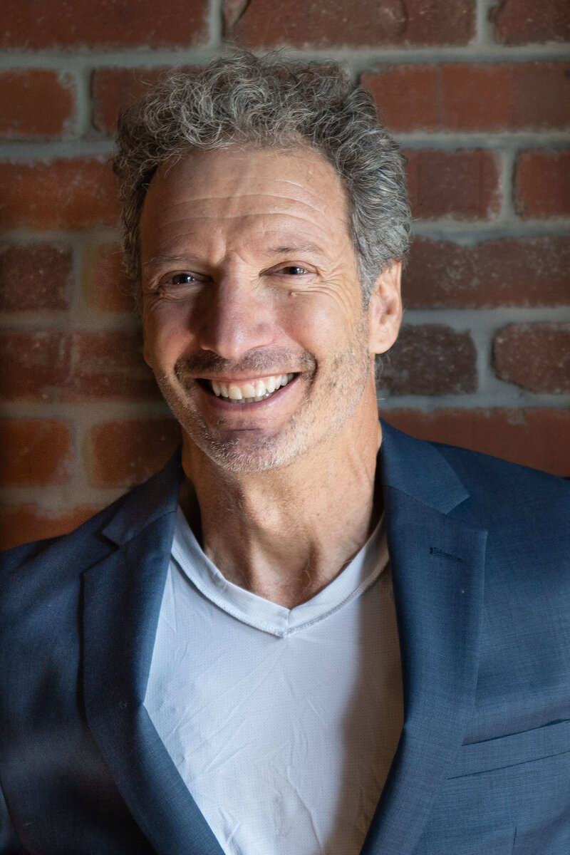 gray haired man in dark suite jacket standing in front of a brick wall