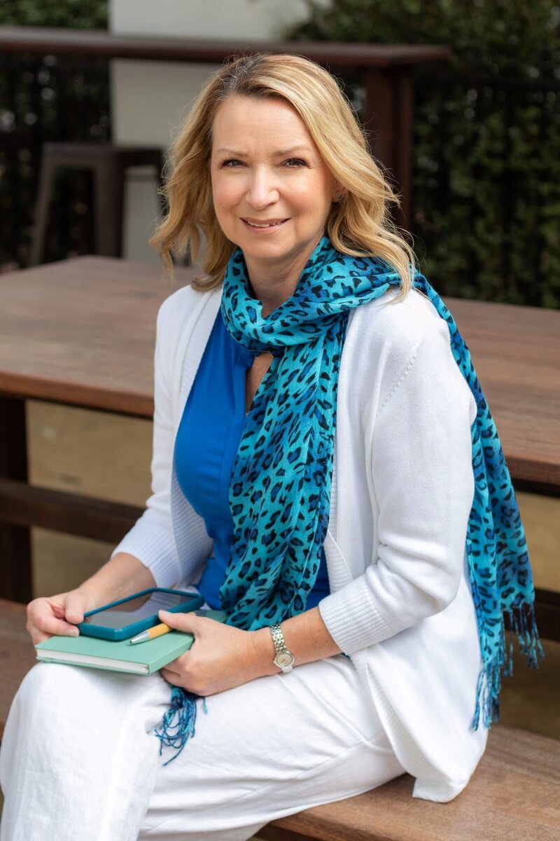 Francine in blue shirt and scarf with white pants and cardigan holding notebook and cell phone smiling at camera