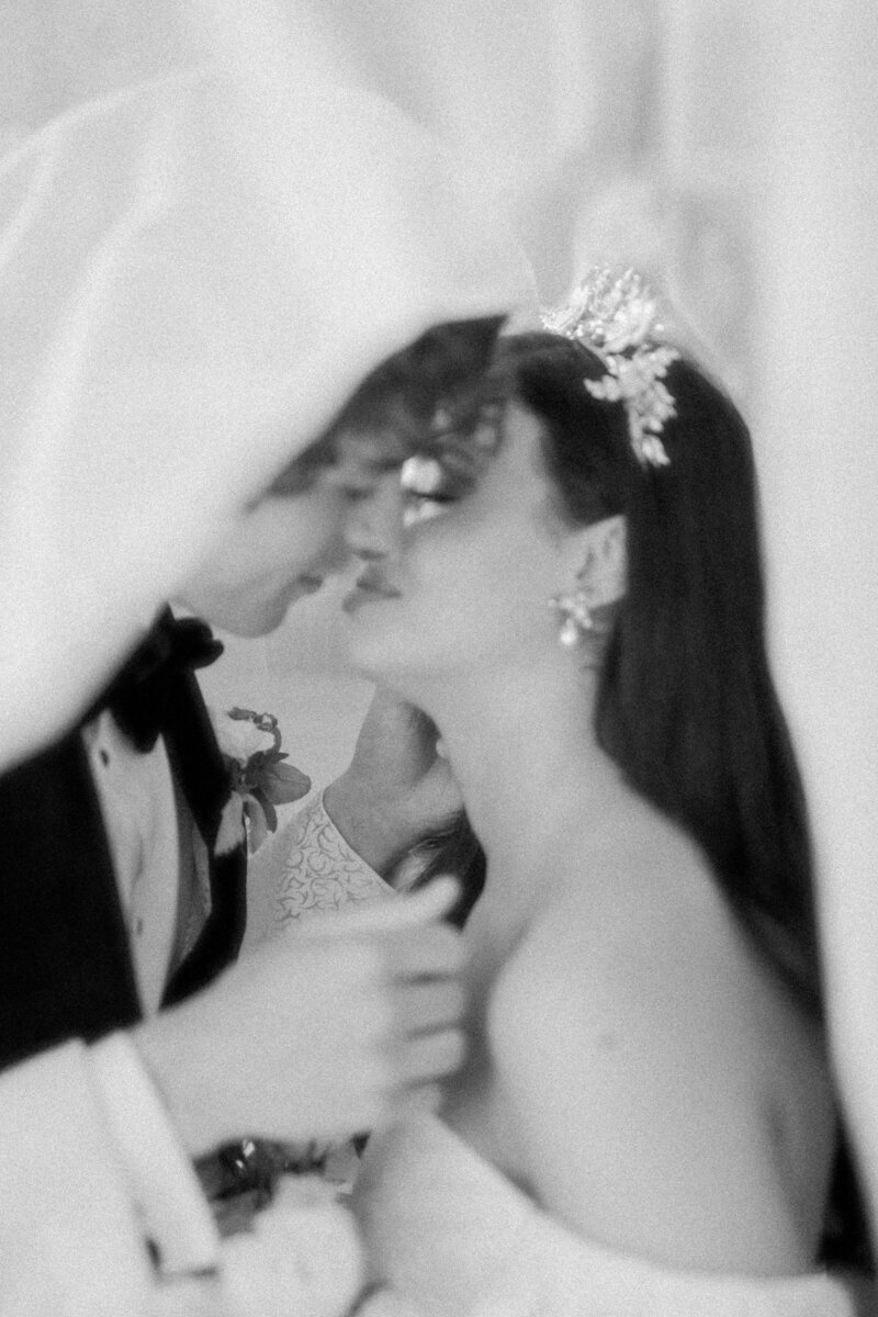 black and white photo of bride and groom under veil kissing