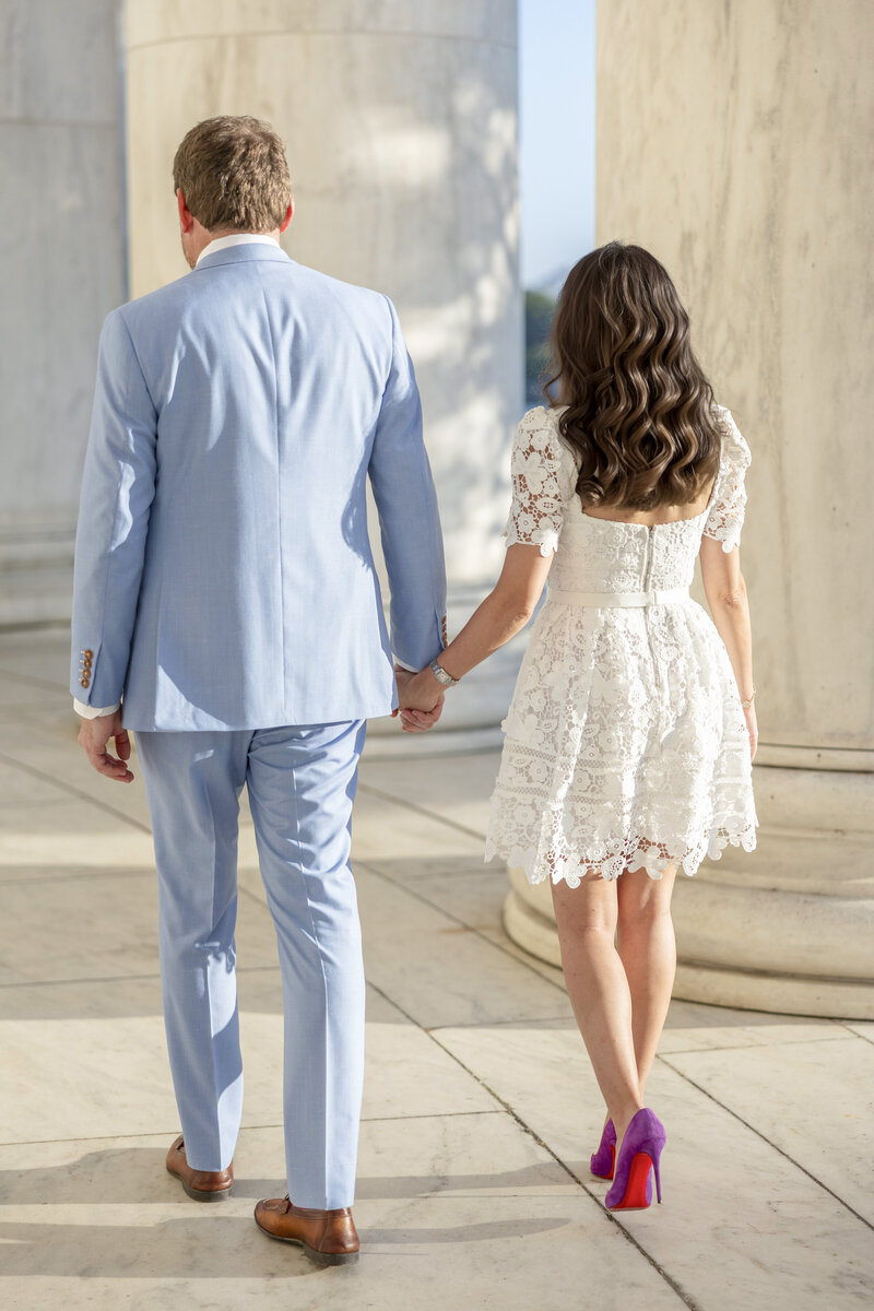 A couple holding hands walks away, with the man in a light blue suit and the woman in a white lace dress and purple heels. They are surrounded by large marble columns.