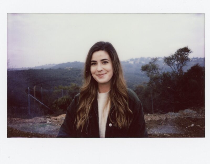 Polaroid of woman wearing dark cardigan smiling at camera in front of green hills