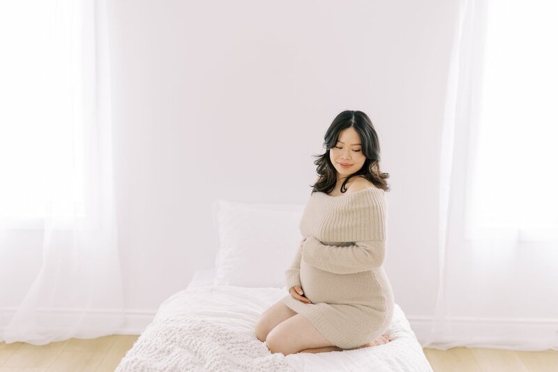 An image of an asian woman wearing a cream coloured dress holding her belly lovingly and looking down as she sits on a white coloured bed in the light bright studio with light coloured flooring