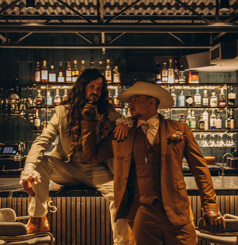 A person sitting on a bar with their partner standing next to them reaching up towards their chin.