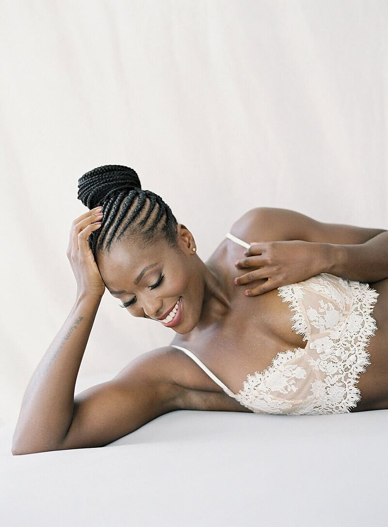 Black woman in cream lace lingerie set smiling down in all white studio with photographer Jacqueline Benet