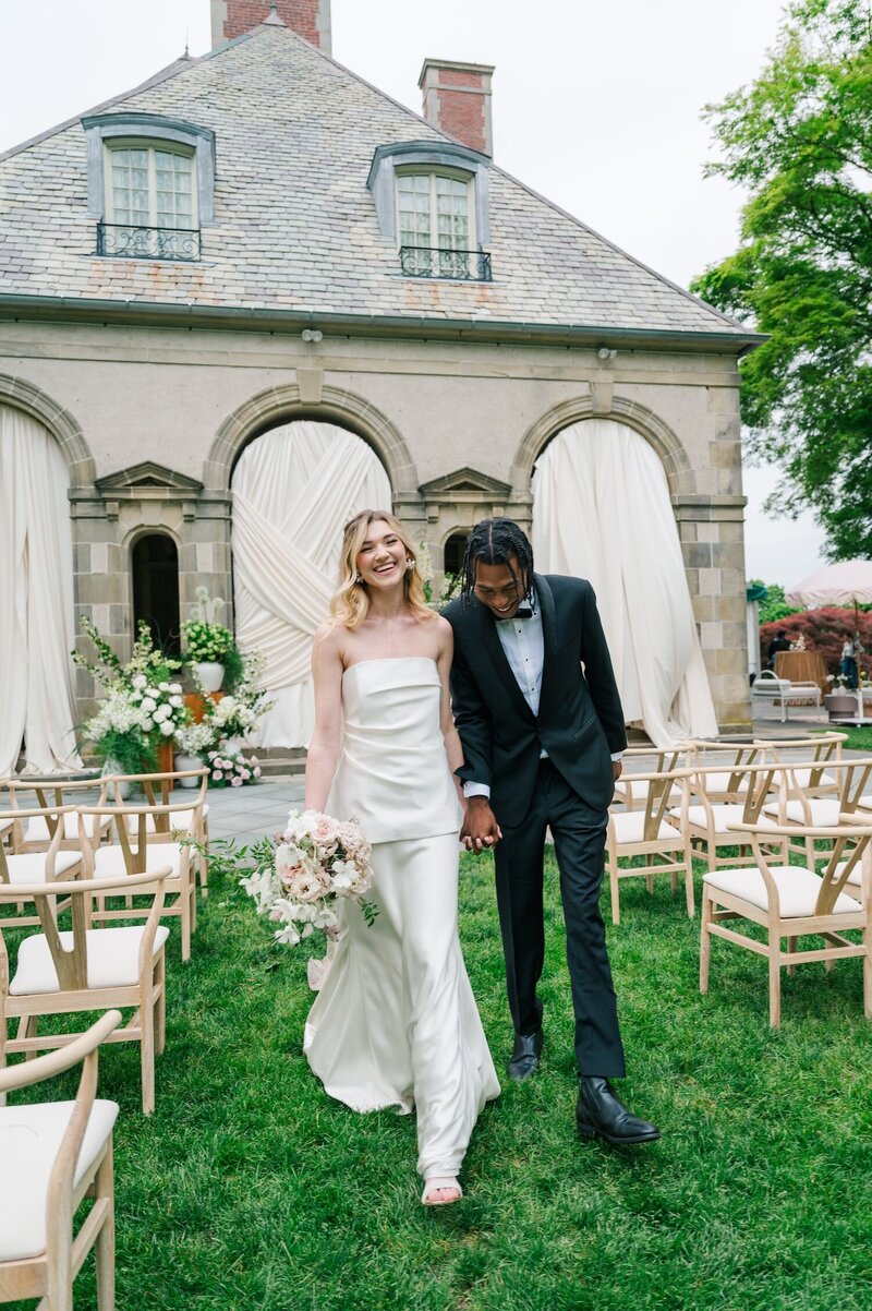 A bride and groom holding hands and laughing as they walk down their ceremony aisle. 
