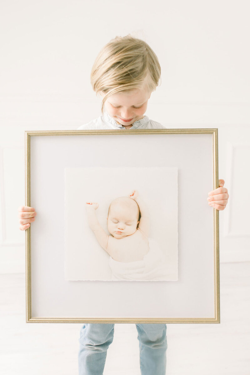 Kleiner Junge hält lächelnd ein Bild mit schmalem goldenen großformatigen  Bilderrahmen und einem Babyfoto.