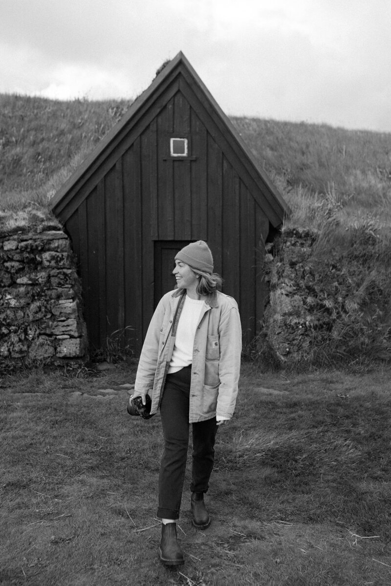 Bare Bones Photo standing with her camera in the Icelandic landscape, surrounded by mountains.
