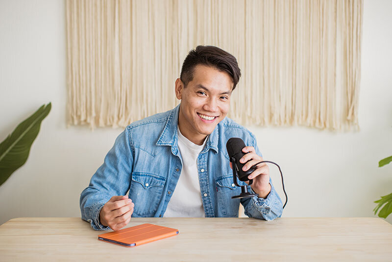 Sho Dewan, young Asian man, career coach and founder of Workhap, wearing a white tshirt and jean jacket, sitting at a desk and holding a microphone in his hand, looking and smiling directly at the camera