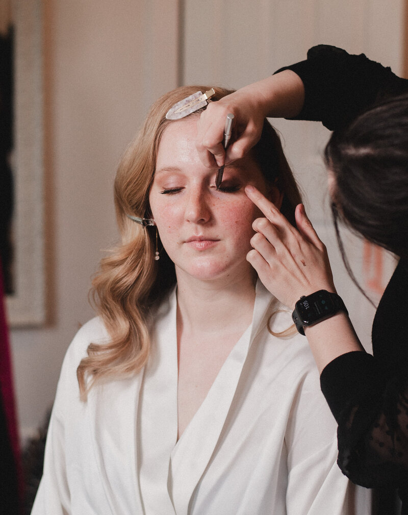 A bride in a robe sits and gets her makeup done by Makeup by Kiley Smith on her Wedding Day