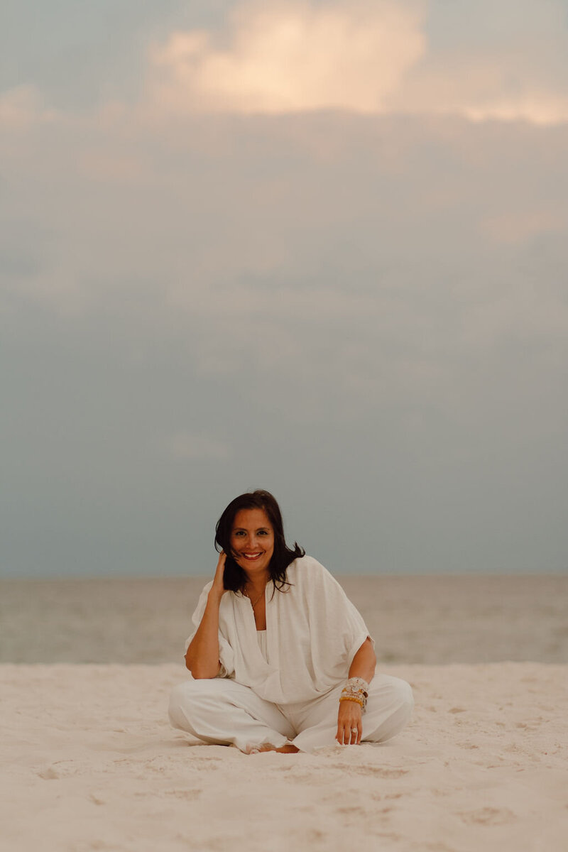 woman with shoulder length dark brown hair smiling, sitting on a beach crossed legged with white pants and white shirt on wearing multiple bracelets