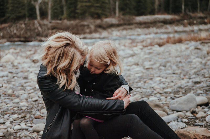 Young mom with blonde hair sits on a mountain creek rock shore looking down at her young daughter in her lap