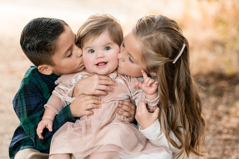 Brother and sister outside holding their baby sister and kissing her cheeks