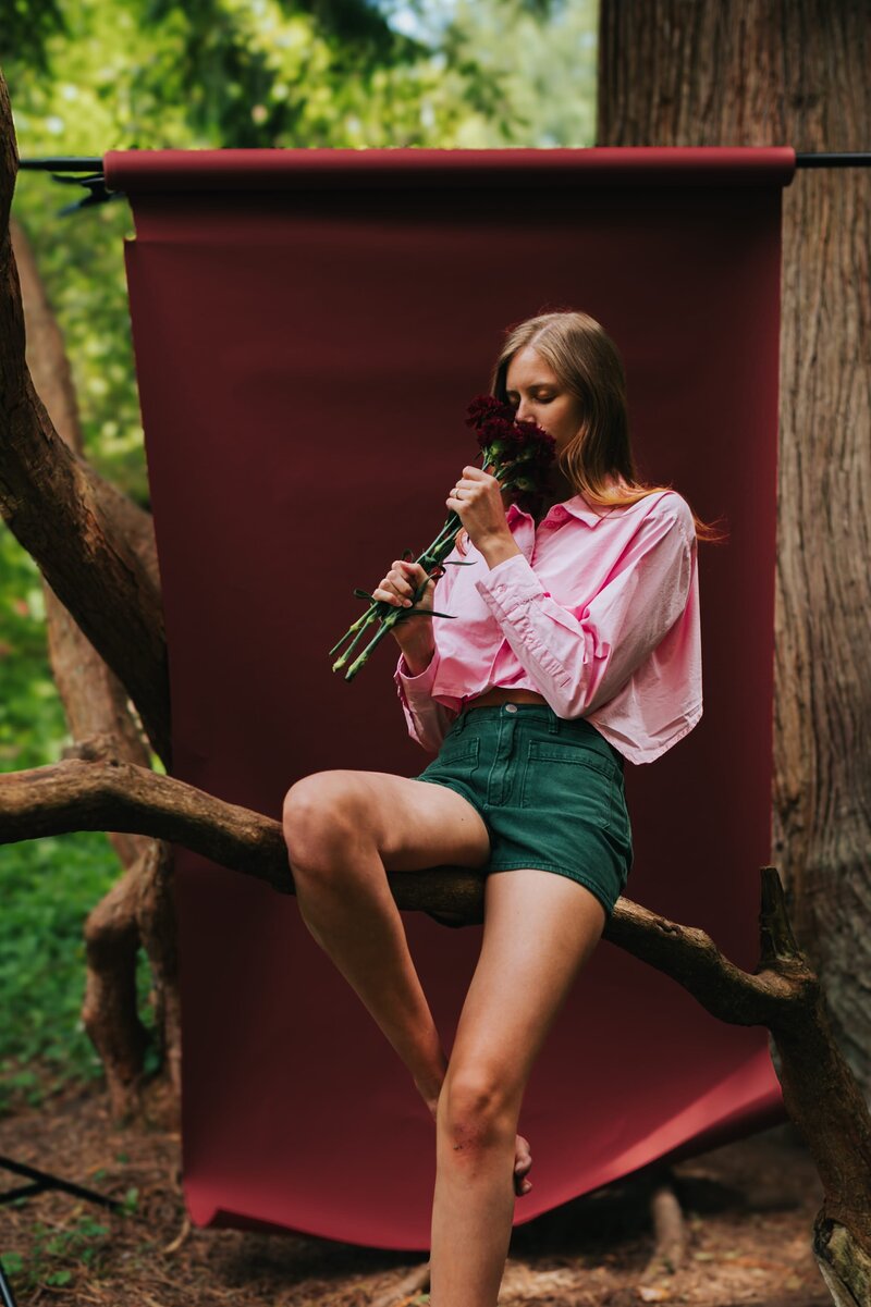 Woman sits on tree branch outdoors in front of burgundy backdrop wearing a pink button down and green shorts with her eyes closed as she smells a bundle of flowers.