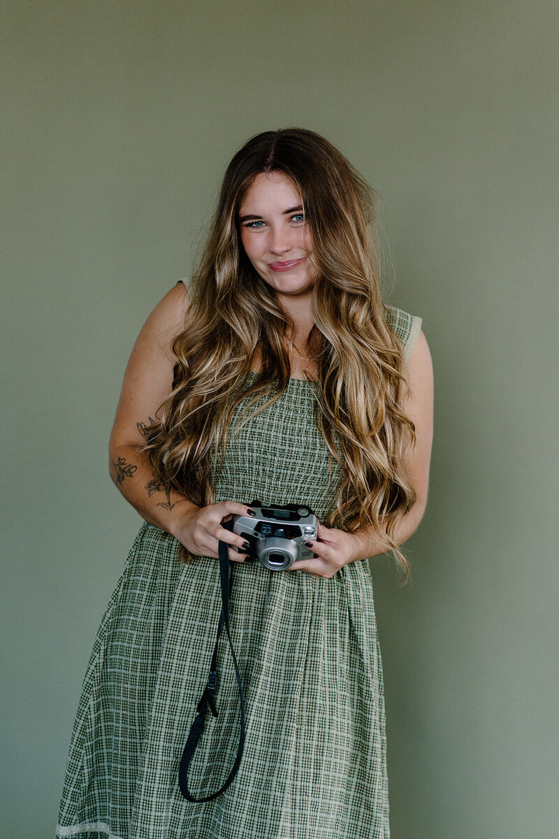 Minnesota wedding photographer wearing green gingham dress holds Canon camera and poses against green backdrop