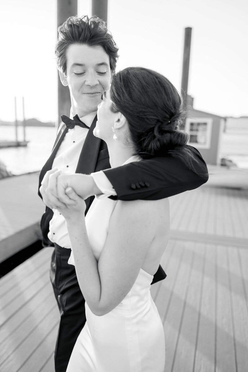 Rhode Island Wedding Photographer | Black and white photo of a groom in a tuxedo and bride in a sleeveless dress dancing joyfully on a wooden dock, with a calm water backdrop.