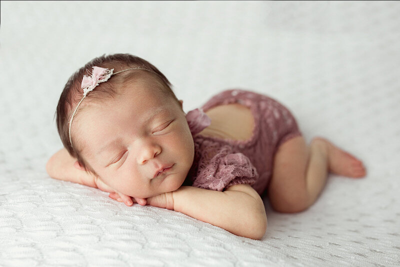 newborn baby girl laying on her belly with her hands under her chin in a pink lace outfit on a white backdrop sleeping peacefully during her harrisburg newborn session