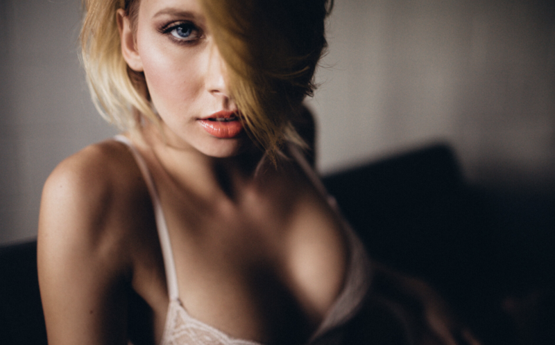 Woman with short blonde hair, poses confidently against a light brick background, wearing a white top. Soft lighting creates a serene mood.