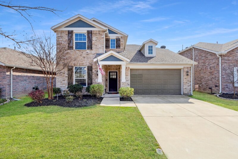 Front exterior of 1708 Ridge Creek Ln, Aubrey, TX featuring a charming two-story brick facade, gabled rooflines, covered porch with American flag, and attached two-car garage — listed by Haven Realty Group.