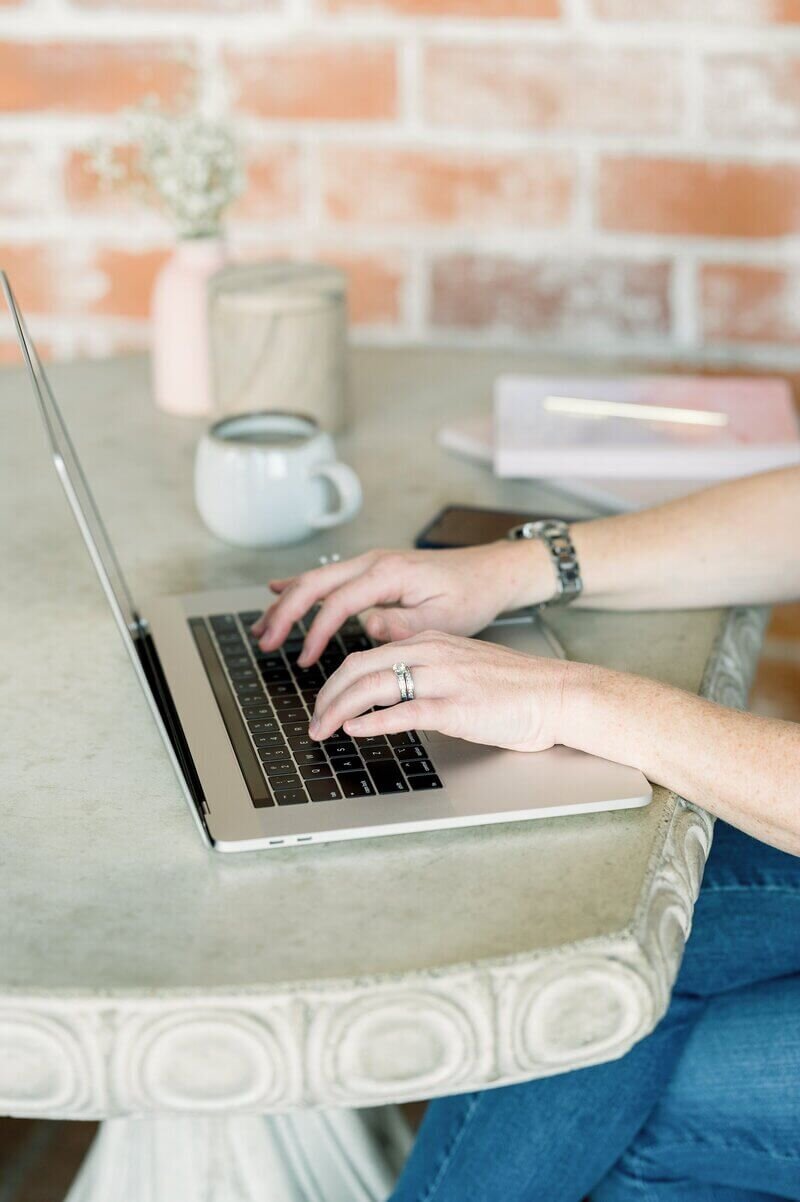 A person types on a laptop at a round stone table with a coffee mug, notebook, and phone nearby, set against a brick wall background.