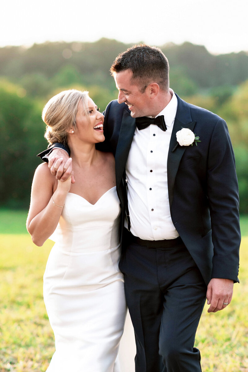 A couple in wedding attire smiles joyfully at each other while walking through a grassy area. The groom wears a black tuxedo with a bow tie and white rose boutonniere, and the bride is in a strapless white gown with her hair in an updo.
