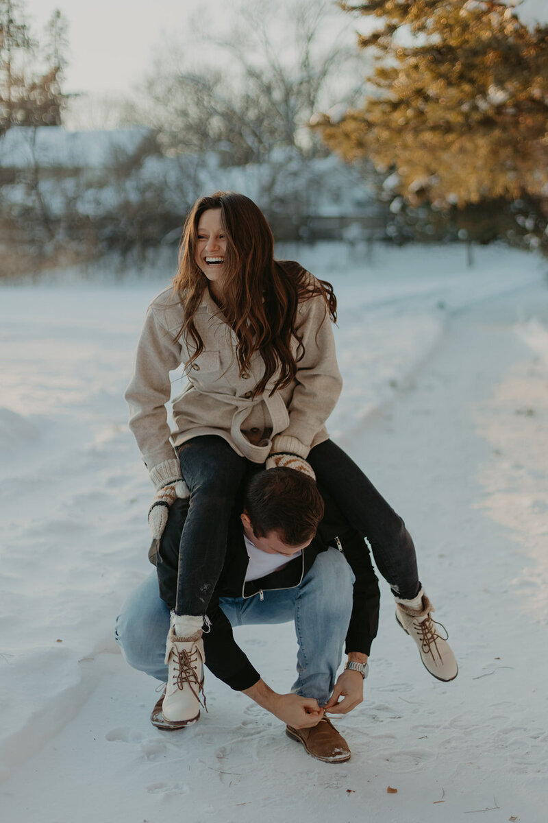 woman on mans back in snow as he ties his shoe