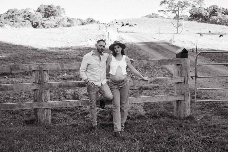Byron Bay Hinterland Maternity photoshoot . A couple lean against a fence in a Pottsville Farm , they look into the camera