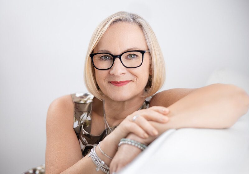 Image of a woman in her 50s on a white background. She is leaning against the sofa for a personal branding style portrait session. Taken by Claire Howes, Norwich.