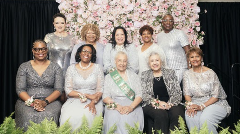 Phi Alpha Omega Charter Members wearing silver gowns in front of a flower wall