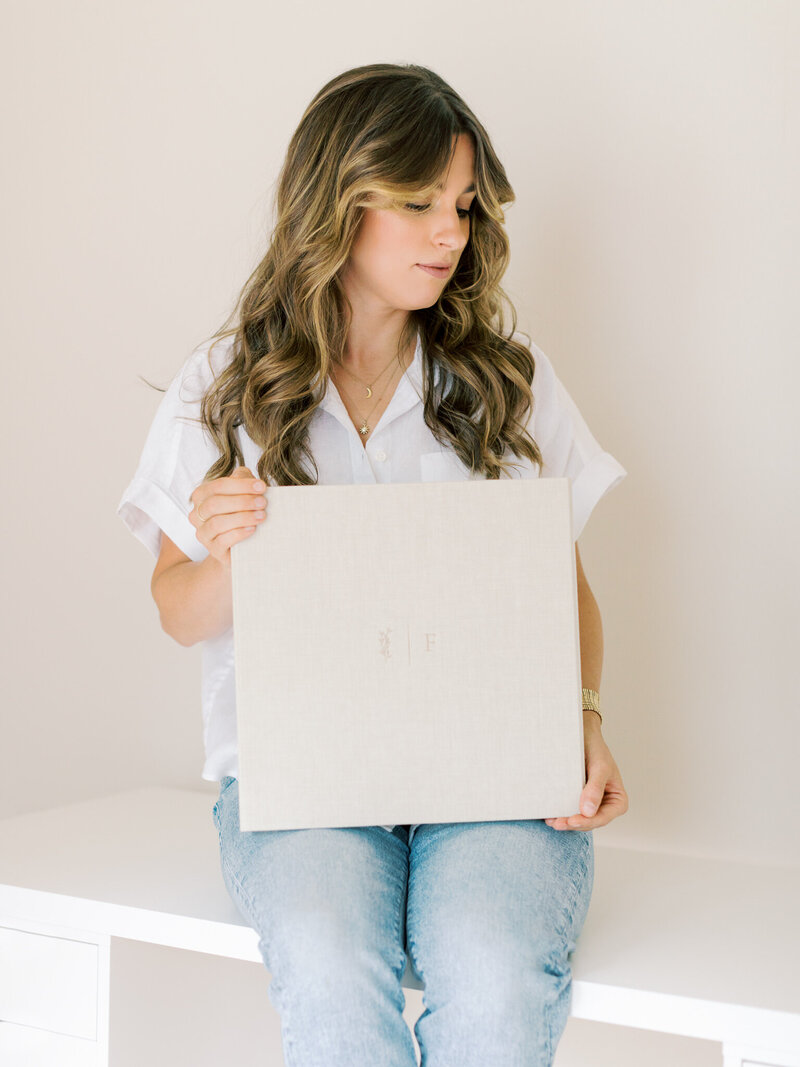 Little Rock Photographer Bailey Feeler sits on white desk holding custom-debossed, linen matted print box