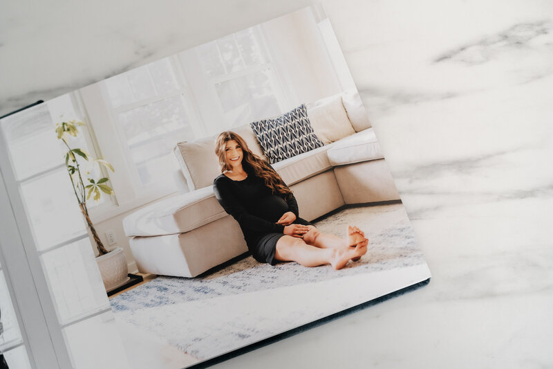 An open photo album featuring a maternity portrait of a woman sitting on the floor in a bright, cozy living room, wearing a black dress and smiling at the camera.