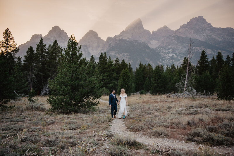 grand teton wedding at dusk in the tetons with a couples walking together on a dirt trail through the woods as the sun shines over the tetons as it gleams through the light fog