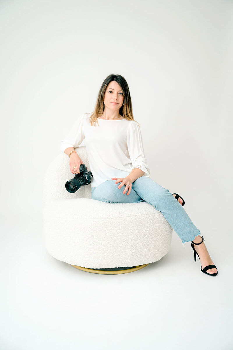 Personal branding portrait of Laura Mariani photographer. She is sitting on a white boucle chair wearing white shirt and jeans with black high heels shoes.