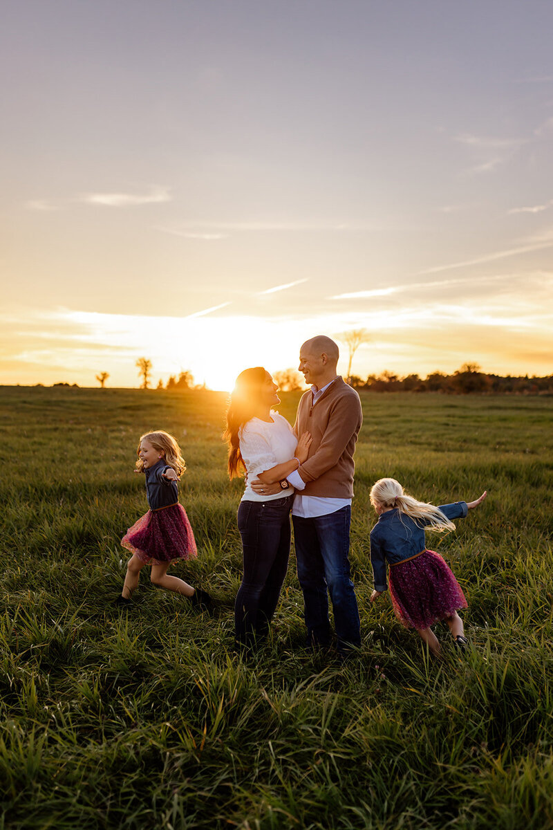 Family photo during sunset at Pinhey's Historic Point in the field with two daughters running around parents who are hugging