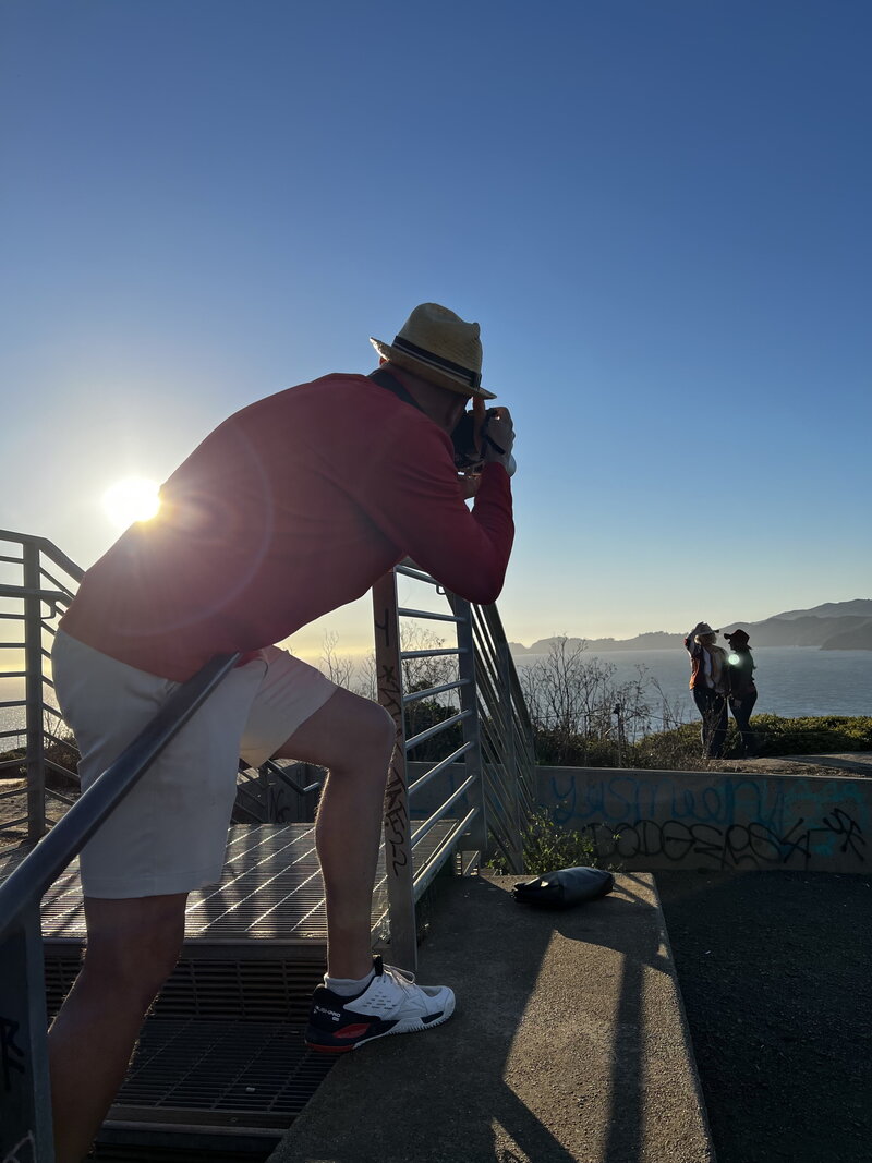 Man leaning on a railing to take a photo of two women at sunset overlooking the ocean