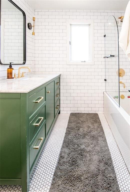 Hall bathroom with white subway tile, black-rimmed mirrors, deep green cabinetry with gold hardware, and tub-shower combo with glass enclosure.