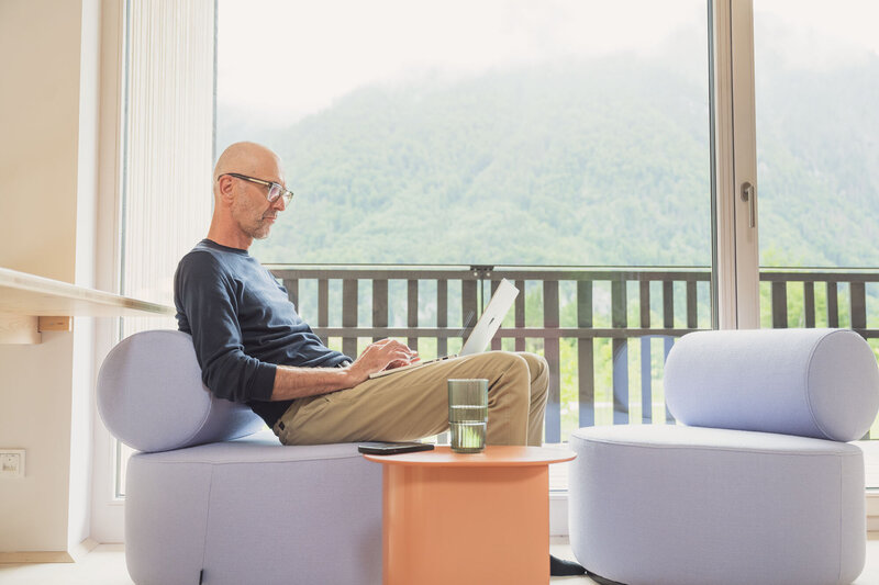 Man working on his laptop with a stunning mountain view, embracing remote work in the Alps. Discover work-and-adventure stays at TheCampusAlps.com.