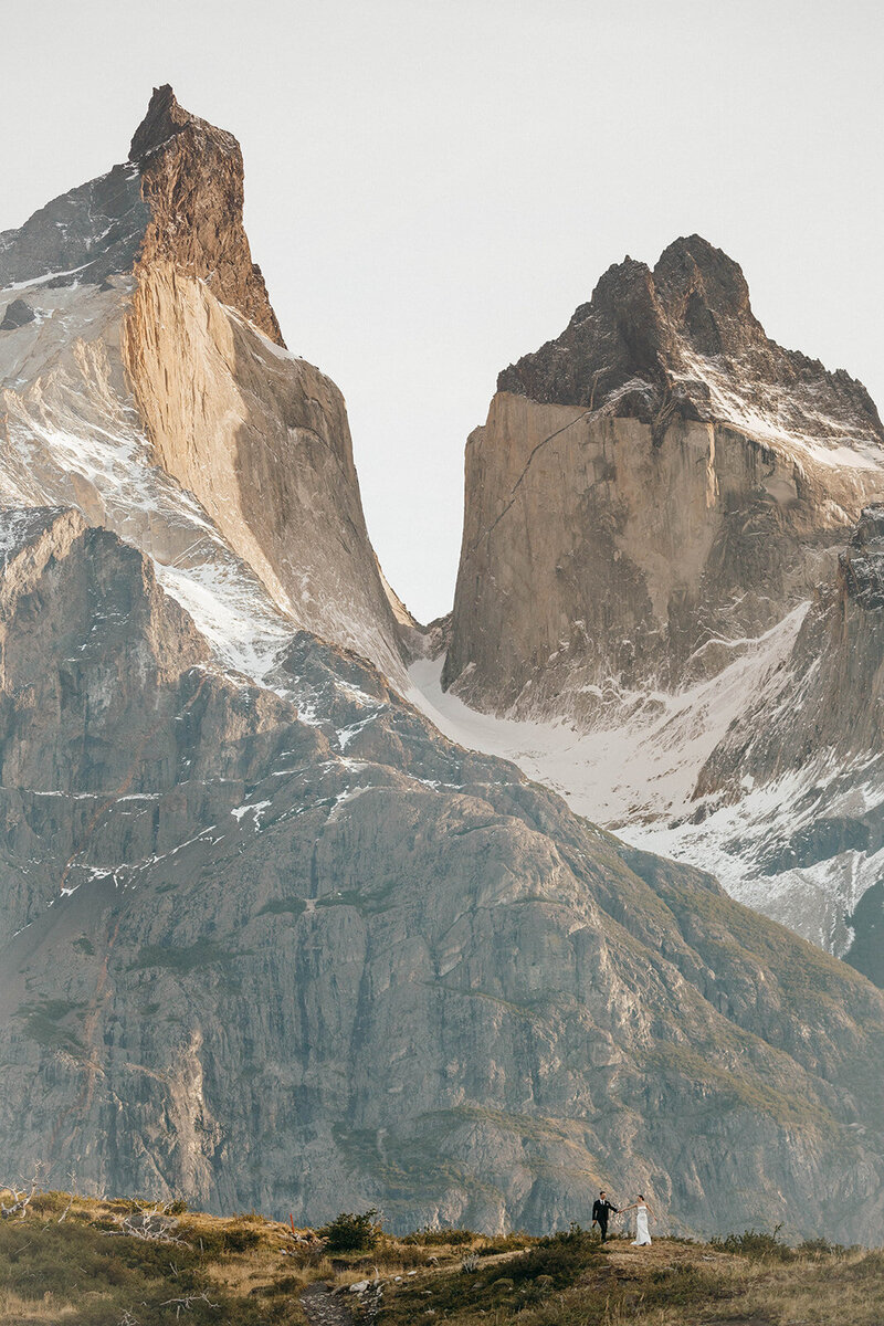 Bride and groom embrace in front of a panoramic view at sunrise with dramatic snow-capped Patagonia mountain peaks in the background.